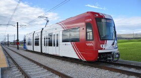 A TRAX light rail train pulls into the station with railroads, green grass, and a blue sky. 