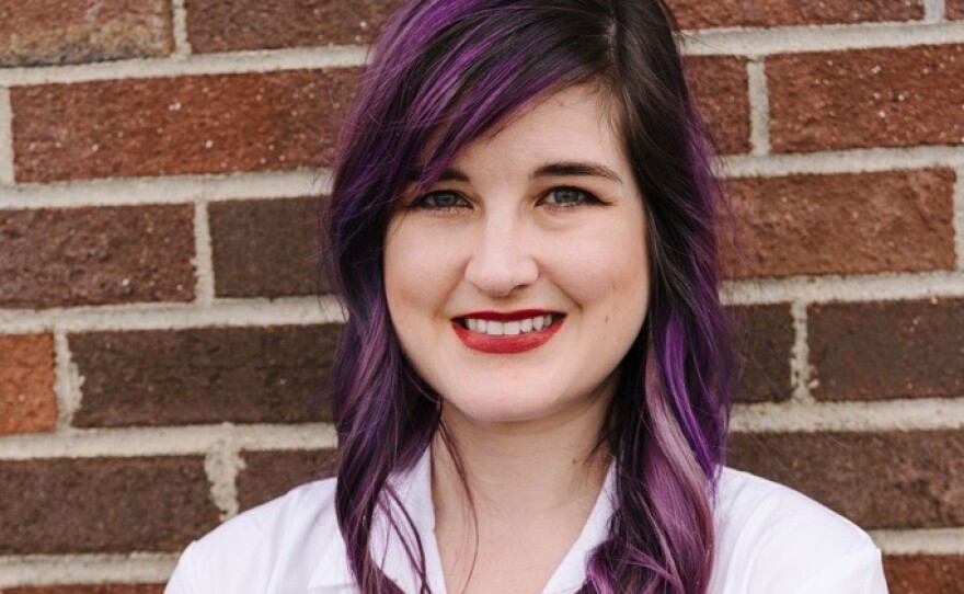 A smiling woman with long purple hair stands against a brick wall with her arms crossed. She is wearing a white lab coat.