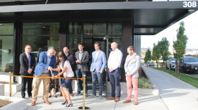 Project leaders stand in front of a new gray and white apartment complex, cutting a ribbon in front of an open entrance. 