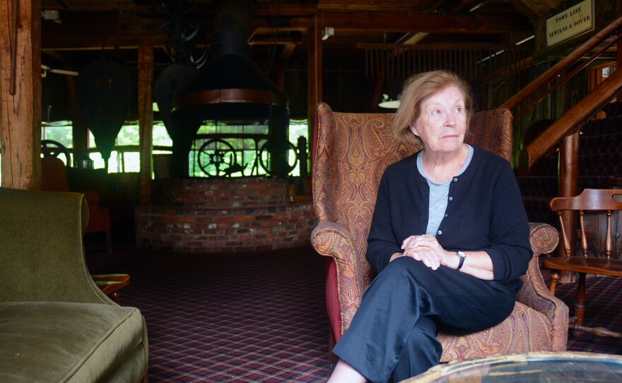 A person sits in a chair with legs crossed and some furniture in the background.