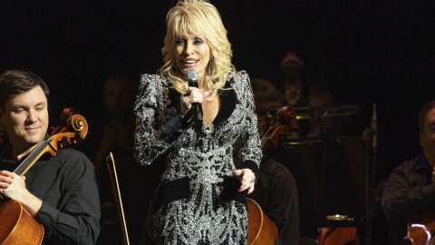Dolly Parton stands on stage holding a microphone wearing a bedazzled outfit. 