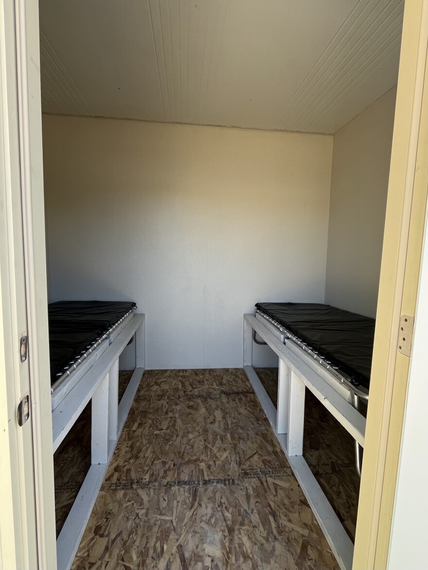 A room in a shipping container, including two black cots with storage underneath. 