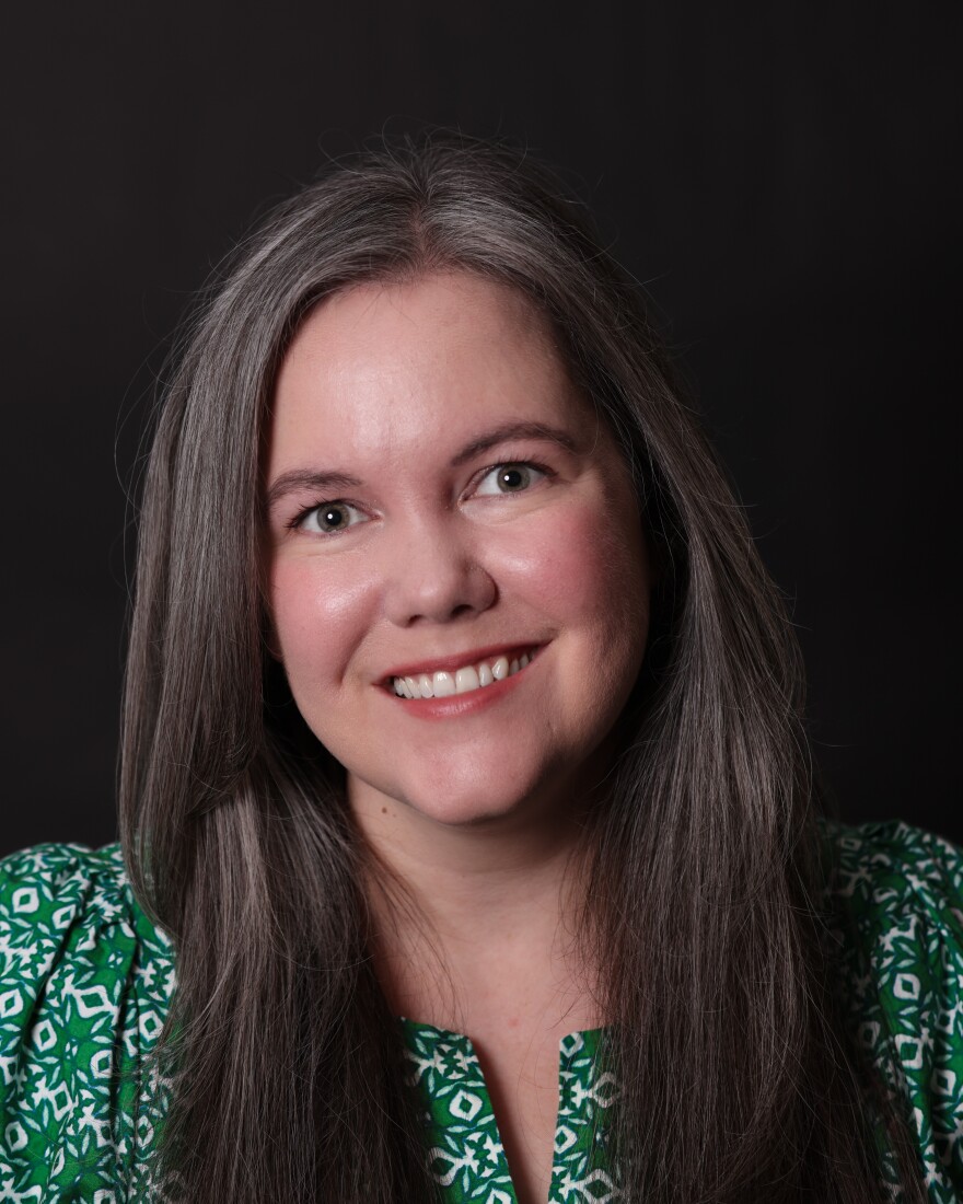A rosy-cheeked woman with brownish-gray hair and a bright smile strikes a professional pose for the camera in a green and white dress. 