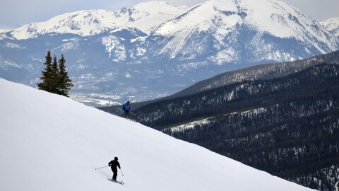 A pair of skiers descends a snowy slope with a view of snow-covered mountains in the distance.