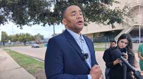 A man wearing a navy blue suit and blue and white shirt is interviewed by reporters. 