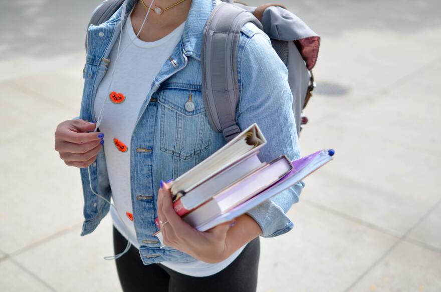 A young woman in a denim jacket holds a binder, books, and a notepad. 