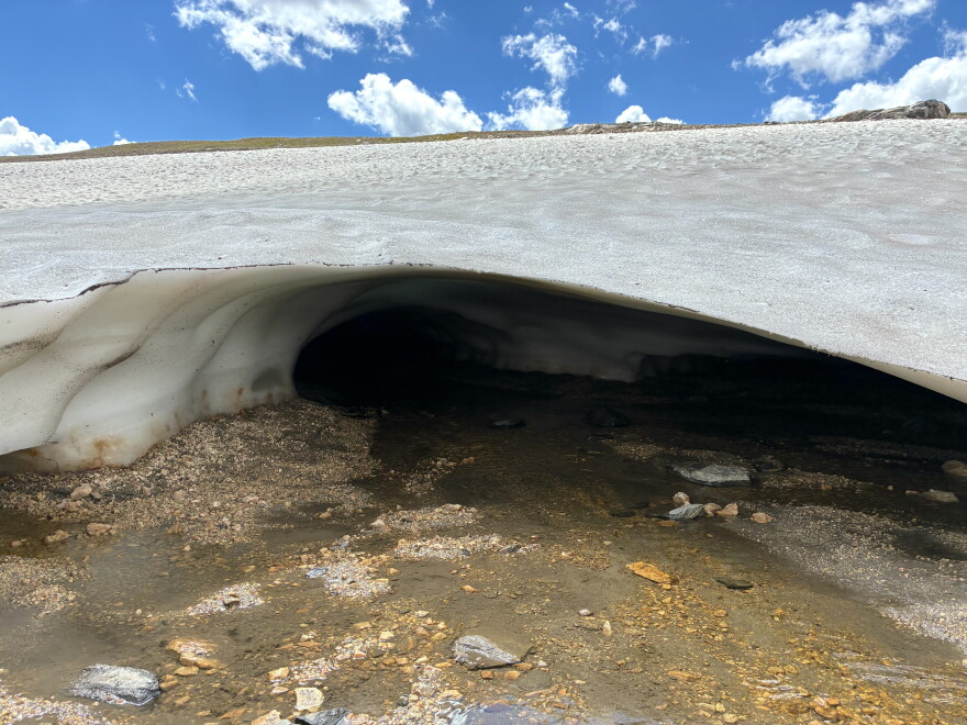 A dark opening beneath an ice patch reveals meltwater flowing across rocks underneath the ice.