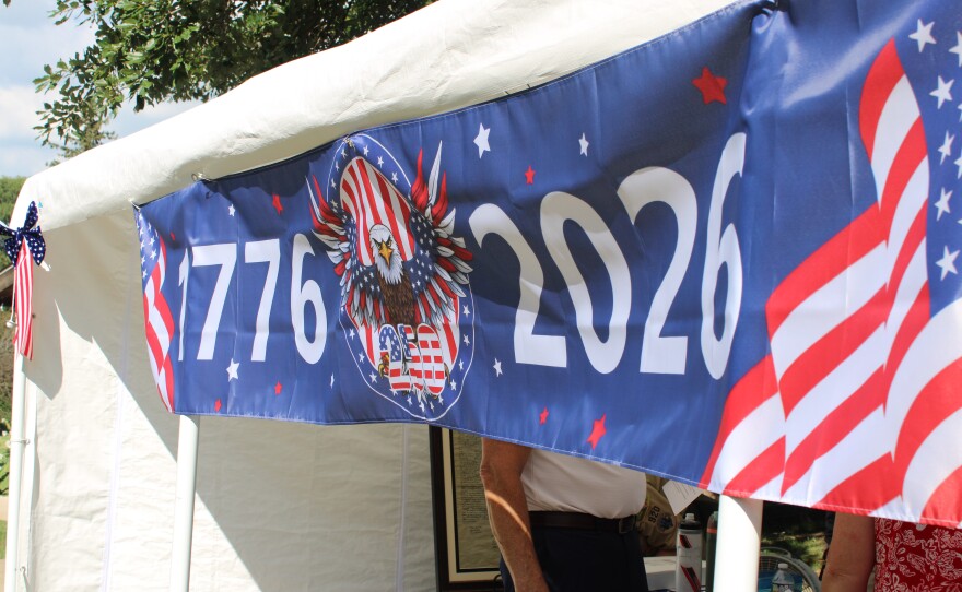 A close-up of a blue banner reading "1776" and "2026" with a bald eagle graphic, hanging from a tent alongside an American flag ribbon.
