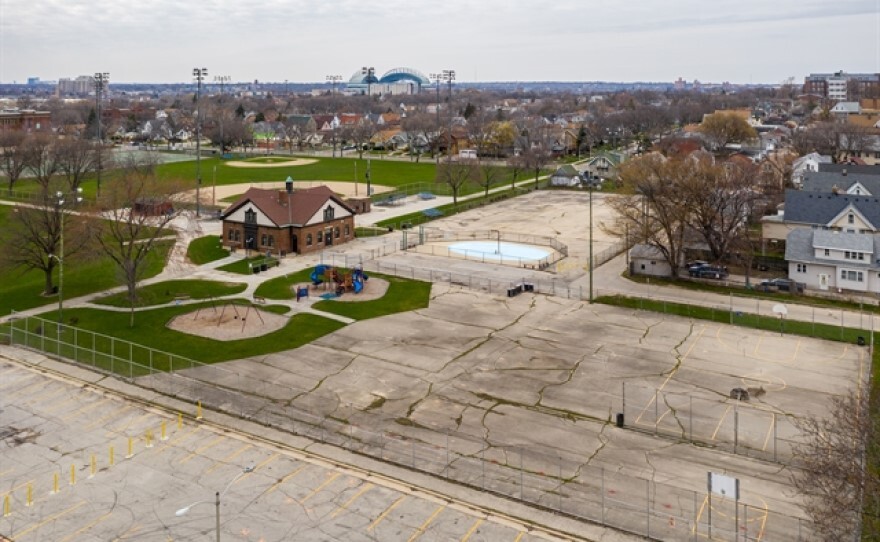 An aerial view of a rundown playfield includes cracked concrete basketball courts and outdated playground equipment.