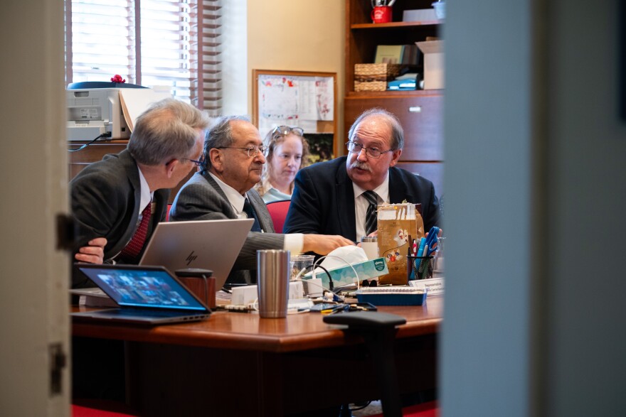 Viewed through an open door, three men wearing suits sit at a cluttered committee table