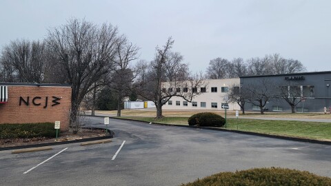A light brick building with black lettering can  be seen in the photo to the left. A two-story building that is gray in the front and white in the back is in the background. A parking lot and side street separate the two buildings.