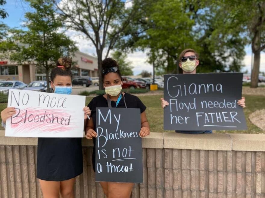 Three people holding protest signs. All wearing masks. The middle reads "My blackness is not a threat."