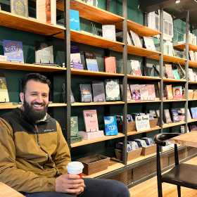 Ahmed Alwhysee poses seated in front of bookshelves with a to-go coffee cup in hand.