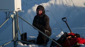 A person bundled up in a snow suit smiles while crouching next to a weather station atop a mountain.