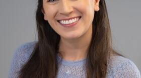 A woman wearing a black sweater sits smiling for the camera on a gray background.