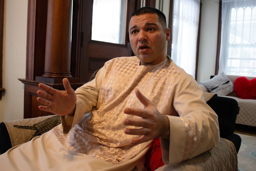 A man in a light colored tunic speaks, gesturing with both hands, while sitting in an armchair.
