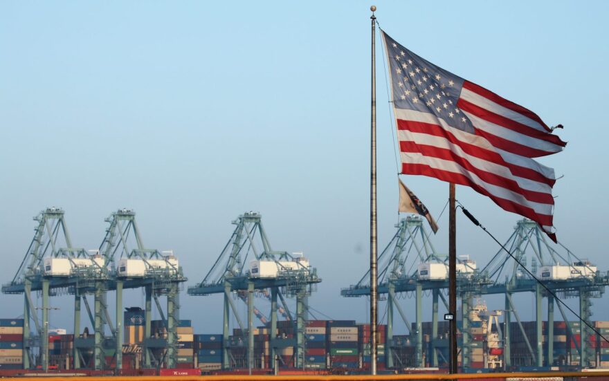 An American flag flies nearby with shipping containers stacked at the Port of Los Angeles in the background, which is the nation's busiest container port.