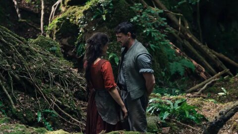 A still from a scene in Hamnet. Jesse Buckley in a red Renaissance dress, stands near Paul Mescal in an English forest. 