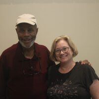 A Black man stands next to a white woman and he has his arm around her shoulders. They're both smiling.