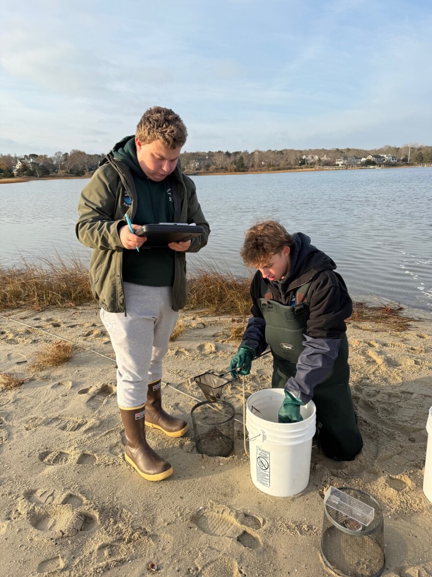 A student with a clipboard stands beside a student looking into a five gallon bucket