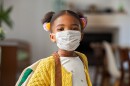 A young girl in a yellow sweater, her hair in pigtails, wears a face mask.