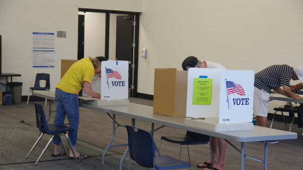 a man and woman fill in a ballot behind a screen that includes an American flag