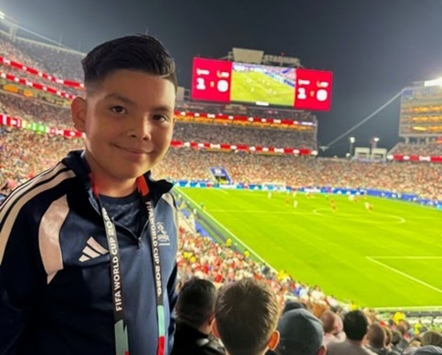 A nine-year-old boy smiles at the camera with a crowded world cup stadium behind him.