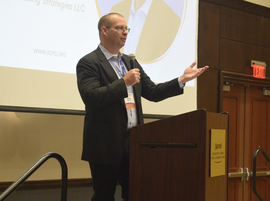 Man in a sports jacket and holding a microphone while speaking at a conference 