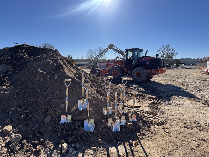 Nine shovels are arrayed in a row in a large pile of dirt. The pile is on a construction site, and a large front loader and an earth drill are parked behind the dirt.