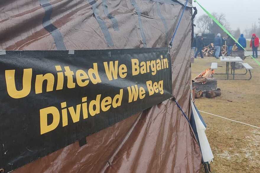 A black tarp-sign attached to a tarp tent reads in yellow lettering  "united we bargain, divided we beg." People can be seen piling firewood on grass in the distant background.