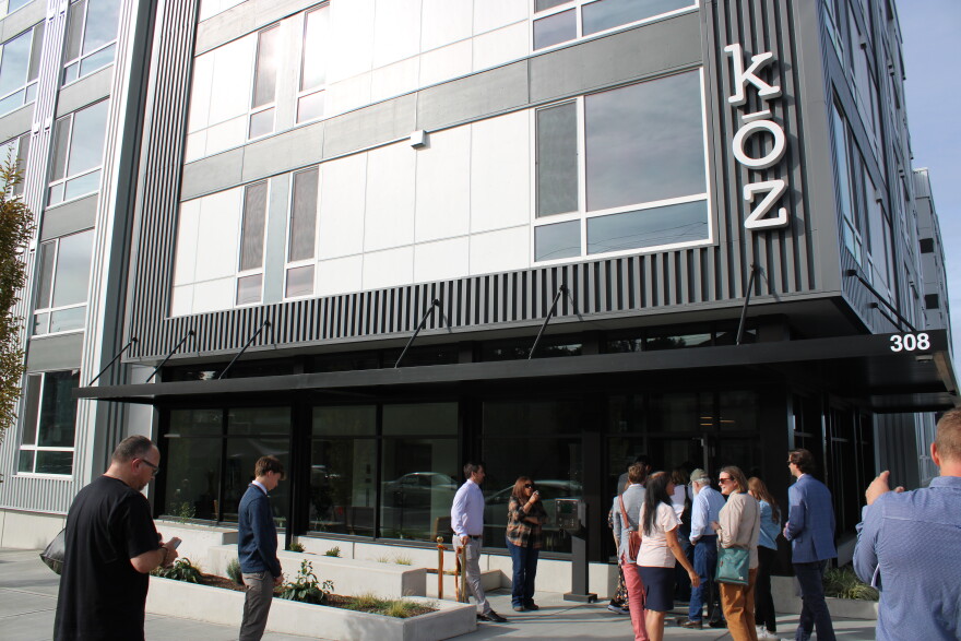 Project leaders and stakeholders stand in front of the apartment complex. Letters in white spell out "Kōz" on the right side of the building. 