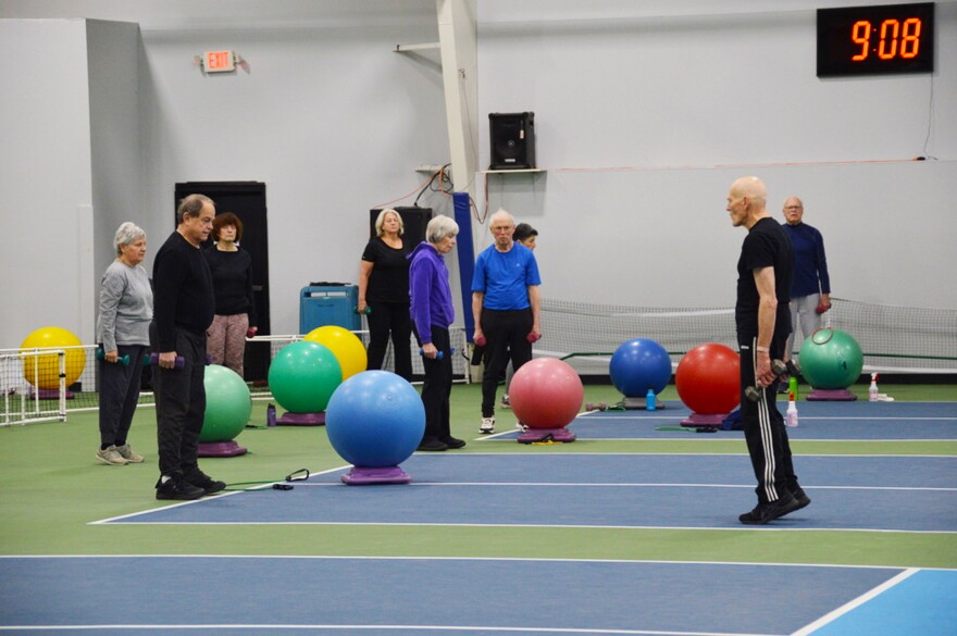 A photo of older people standing in a gym space holding small weights. Large colorful balls are spread throughout the people, too. 