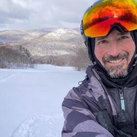 Man with beard and ski helmet standing on ski slopes with mountains in distance