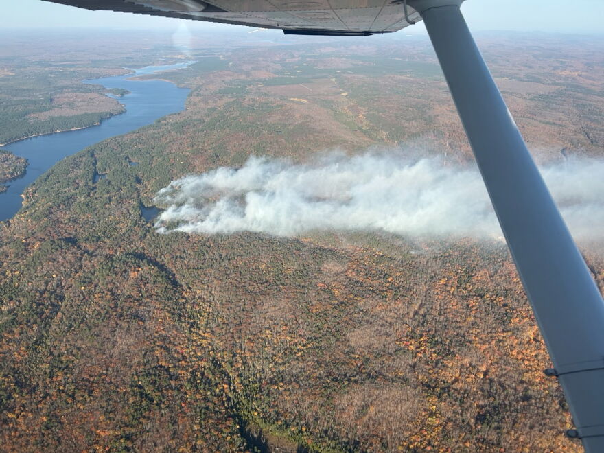 An aerial photograph taken from inside a small aircraft shows a large plume of thick, white smoke rising from a dense forest during autumn. The wing strut of the airplane is visible on the right side of the frame. Below, a sprawling landscape of trees shows changing fall colors, with shades of orange, yellow, and brown mixed with green evergreens. 