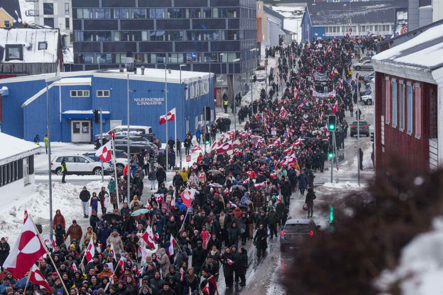 A large crowd marches down a snowy street, many holding Greenland flags. People are bundled in winter clothing, and buildings line both sides of the street under a cloudy sky.