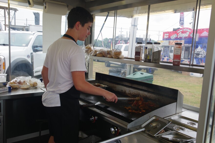 A youth stands inside a small kitchen, a large window in front of the grill he's working at shows another fair concession stand just in front of him. He's using silver prongs to cook an assortment of toppings for food he's preparing for customers. He wears a white t-shirt and black shorts, with a matching black apron. Bags of hamburger buns sit to the right of him and on the silver metal shelf above the griddle. An assortment of spices and other ingredients can also be seen on the shelf. 