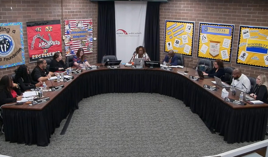 A group of people sit around a large U-shaped table in a meeting room with bulletin boards on the walls and “Peoria Public Schools” on a banner behind the center person.