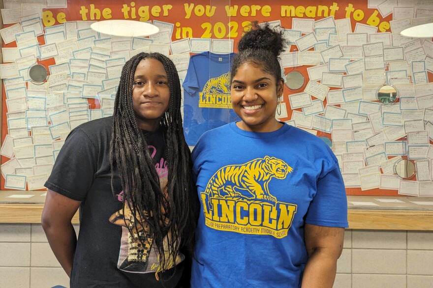 Two people stand in front of a bulletin board in a school hallway. They are smiling and looking the camera.