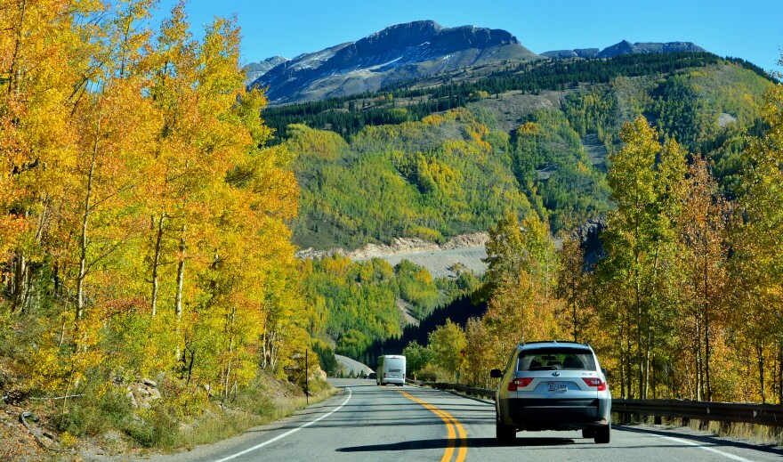 This is an image of a two lane highway in a mountain area with fall foliage lining the sides of the road. There is a silver SUV driving on the road. 
