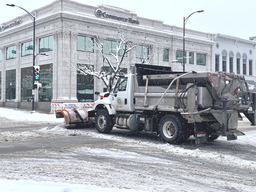The side and back of a City of Columbia snowplow is visible as it pushes snow with its orange blade through an intersection in downtown Columbia.