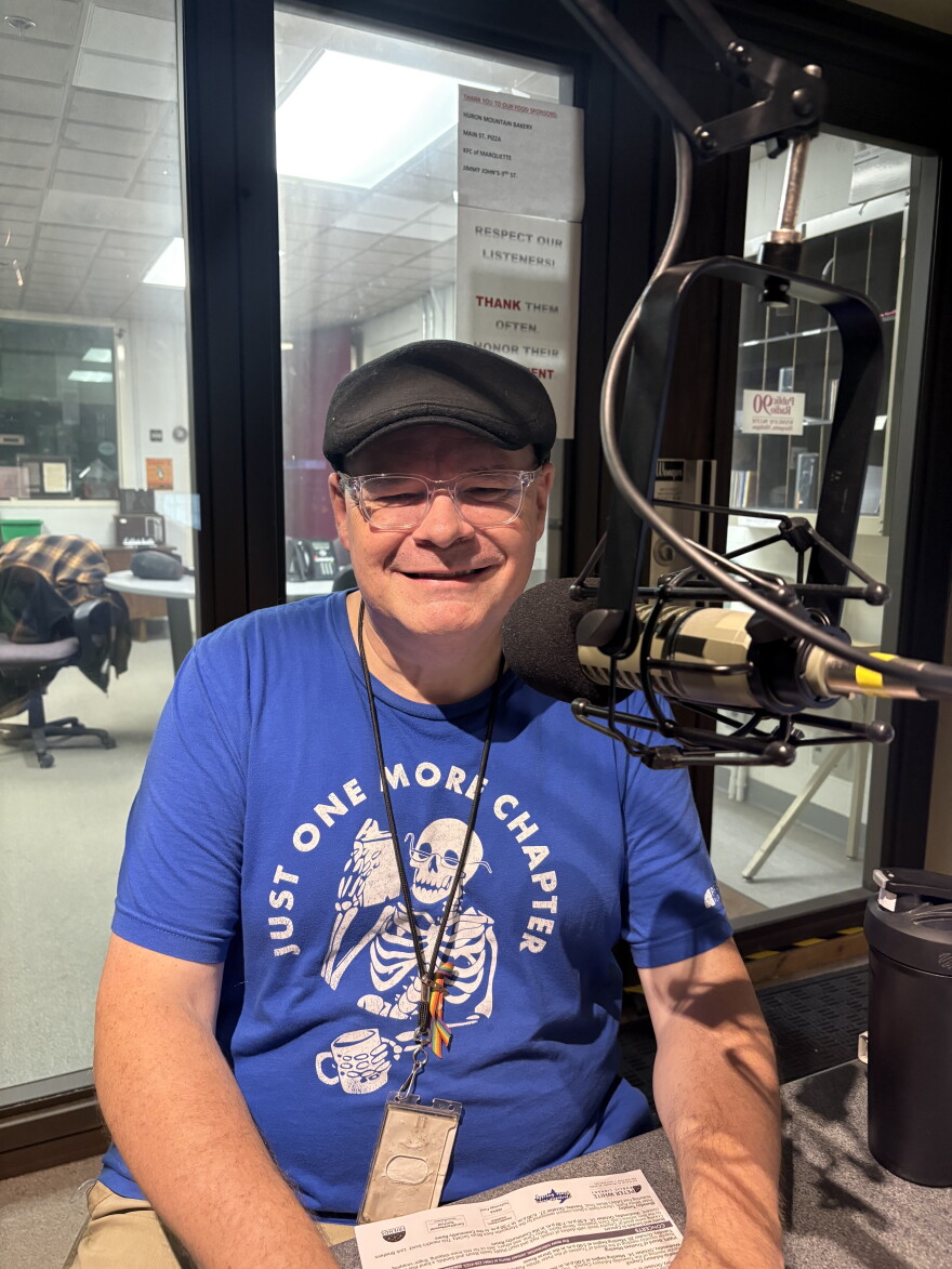 A man sitting and facing forward and smiling near a microphone, wearing a hat and a blue shirt