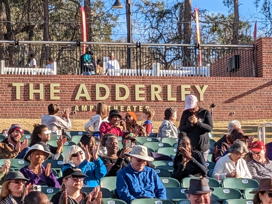 A number of people sit in stands with a red-brick wall with gold letters that read " The Adderley Amphitheater" behind them.