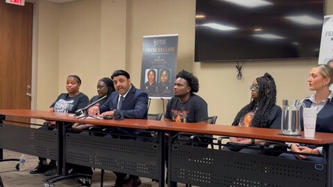 Attorney Shiraz Khan sits by Clark's mother to his right and Clark's son to his left. Large promotional posters from the Shiraz Law Firm bearing the headline "Press Release: Justice for Krystal Clark" stand in the background.