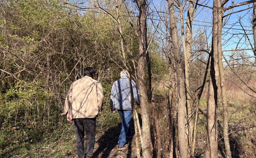 Photo of two people walking away from the camera. They are outdoors in a wooded area. It's very early spring, so there is more brown than green, though some small new leaves are visible on some tree branches.