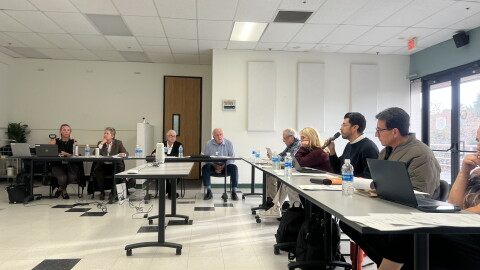 Several people sit a tables in an "L" shape. A speaker on the right holds a microphone.