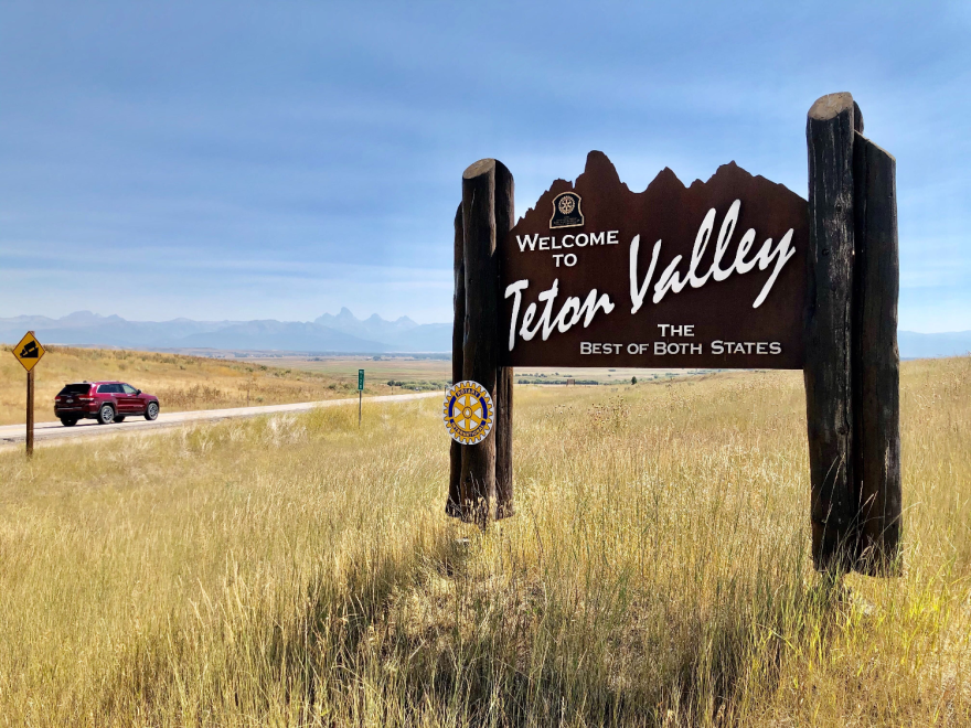 A sign reads "Welcome to Teton Valley, the best of both states" in a flat, grassy area with mountains in the distance and a car driving down the road nearby. The metal sign is cut to look like the same mountains in the background.