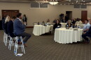 Four individuals sit on stools in front of people sitting at several round tables with white tablecloths. The person closest to the camera, on a stool,  is holding a microphone. 