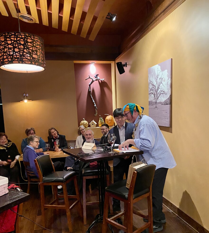 A group of people are seated watching two people speaking at a high-top table in a earth-toned wine bar