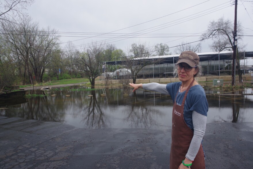 A woman in a blue t-shirt with a long sleeve grey shirt underneath points towards a large pool of water that has gathered in a parking lot. She is also wearing a light brown baseball cap and deep brown apron. The weather is overcast. 