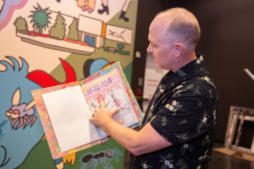 A man wearing a black floral button up shirt stands in front of a colorful mural with an open sketchbook.